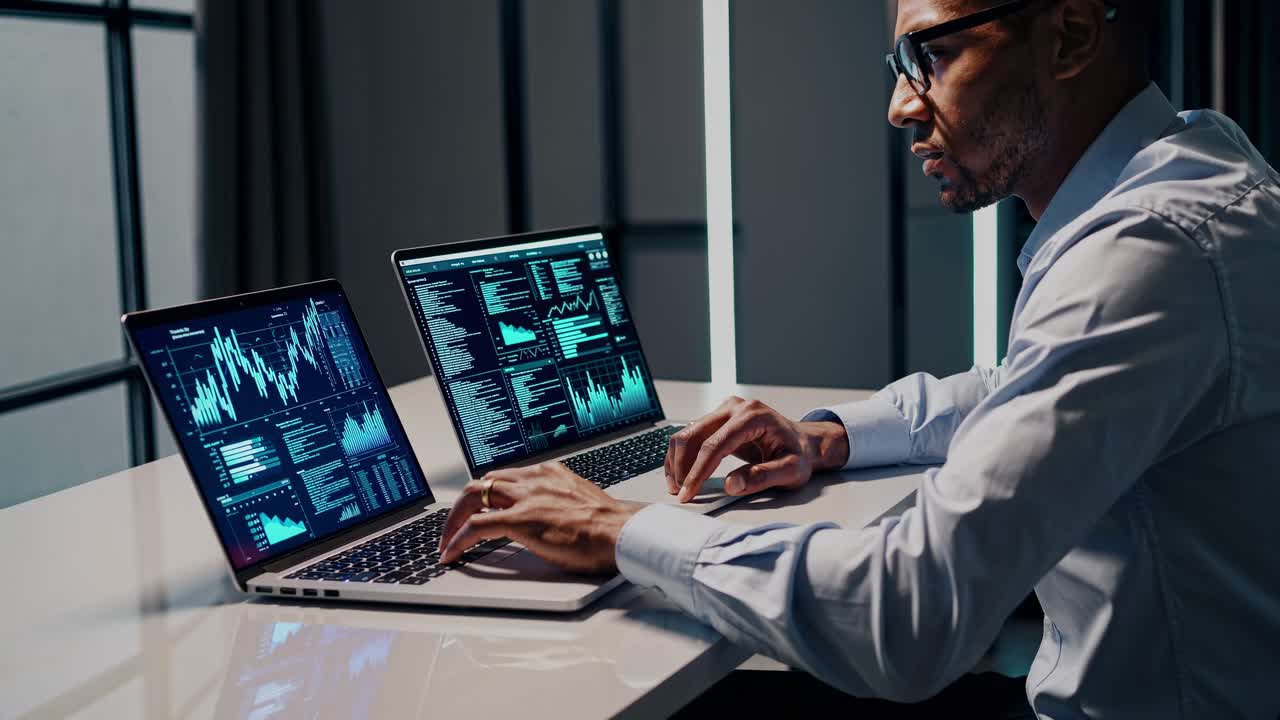 A side-angle video captures a focused professional analyzing data on dual laptops in a modern