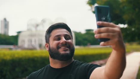 young man smiling and taking a photo selfie at a beautiful park botanical garden with a big greenhouse and garden on the background
