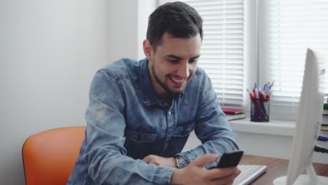 Young-happy-office-worker-using-his-phone-at-the-office-sitting-at-the-table-with-computer,-phone-and-cup-and-smiling.-Shot-in-4k