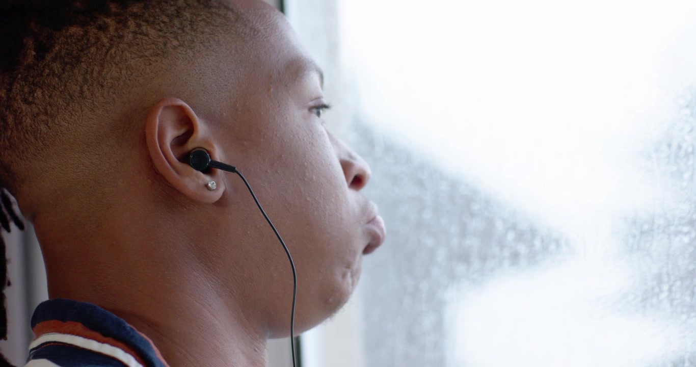 African American man sitting beside wet window striped polo wired buds puffing relaxing, copy space