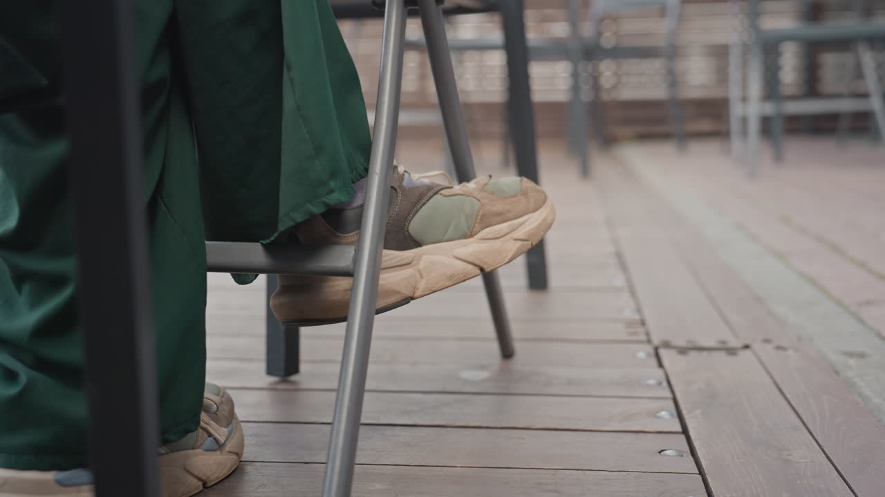 pies sobre la tarima de madera debajo de una mesa. Artista descansando en una silla con zapatillas de esparto, pantalones verdes, patas metálicas de la silla, poca profundidad de campo, ambiente de patio urbano, luz de la mañana, momento de espera tranquilo