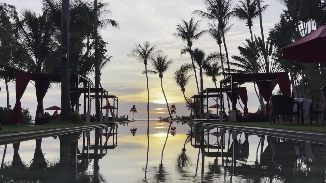 Reflective lap pool leads to a stunning Ko Samui sunset, flanked by red cabanas and palm silhouettes, evoking tropical luxury. The golden sky and tranquil waters offer a dreamy escape
