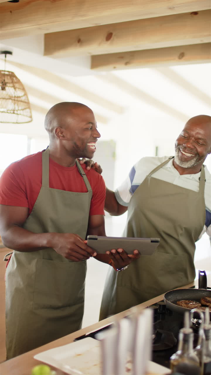 Vertical video: Cooking together, two men in aprons using tablet in kitchen, smiling