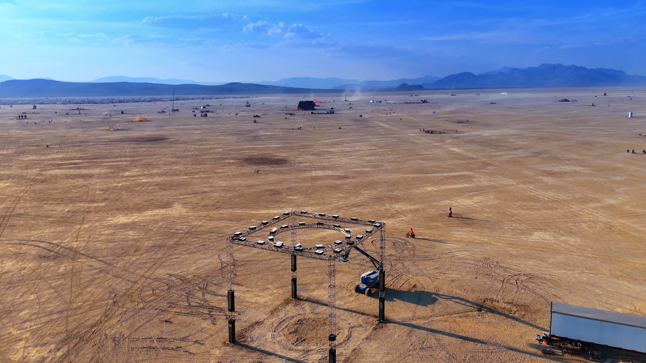 A stage being set up in the vast desert. People walk and ride by the Black Rock Desert. The Burning Man festival from aerial view