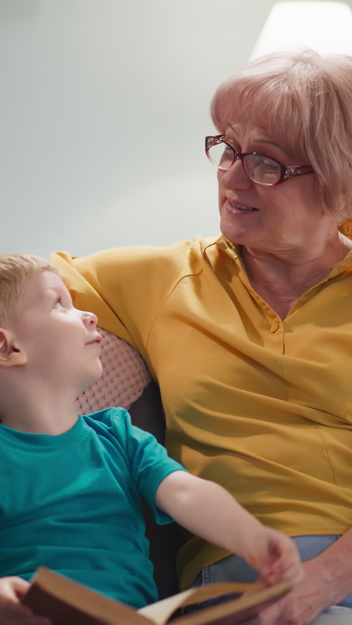 Funny little blond boy turns book pages with granny in living room slow motion. Aged lady in glasses tells story to grandson at home. Woman babysitting child