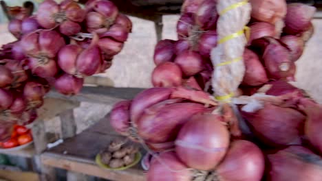 Bunches-of-red-onion-shallots-hanging-from-a-rural-market-stall-with-fresh,-organic-produce-in-Southeast-Asia