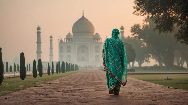 Vue de dos d'une femme en sari vert marchant sur le chemin principal vers le Taj Mahal au lever du soleil à Agra, en Inde, avec une douce brume matinale et des cyprès.