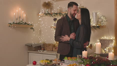 elegant couple standing at christmas dinner table and tenderly embracing each other while woman looking at camera