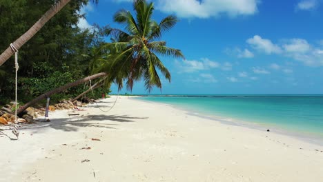 Paradise-beach-with-palm-trees-bent-over-white-sandy-beach-washed-by-turquoise-lagoon-under-bright-blue-sky-with-white-clouds-in-Thailand