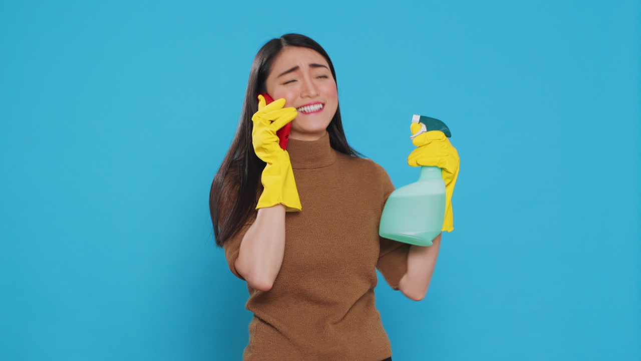 Cheerful housewife wearing protective gloves while holding chemical spray bottle talking at smartphone