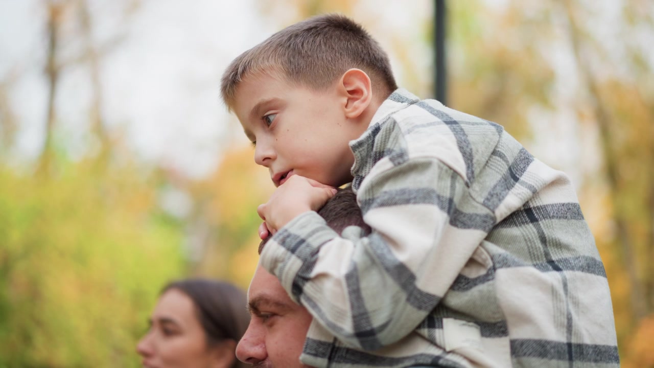 Primo piano di un bambino che riposa sulle spalle di un adulto in un parco all'aperto. Il bambino indossa una camicia a quadri e si tiene delicatamente all'uomo, con alberi visibili e uno sfondo di luce diurna soffusa. L'adulto porta il bambino.