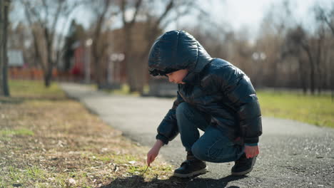 a young boy, dressed in a shiny black jacket, blue jeans, and boots, crouches on a paved path playing with a stick on the ground, with a view of leafless trees blur house view