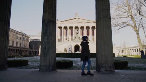 caucasian woman walks in front of tall stone columns with imperial and impressive berlin unesco old national gallery museum building in background, germany, static profile slow motion