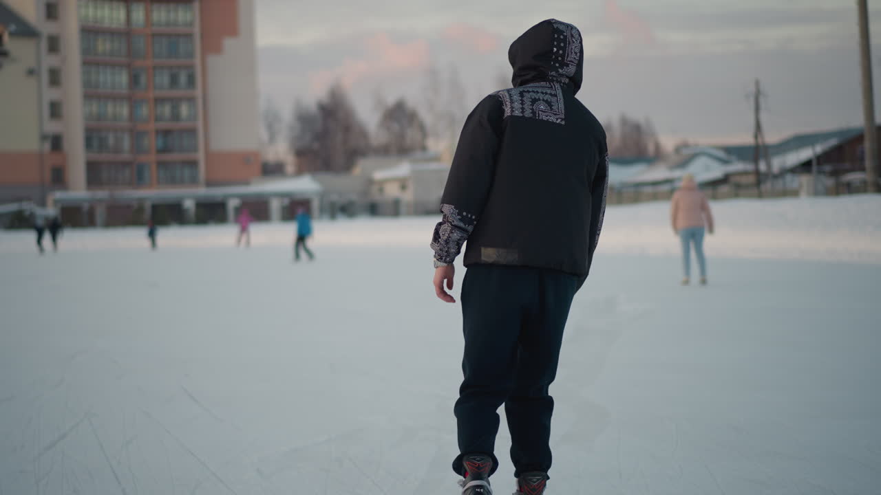 Young people skating on outdoor ice rink during winter season wearing warm clothes, enjoying outdoor activity in winter park with buildings and trees in soft light background