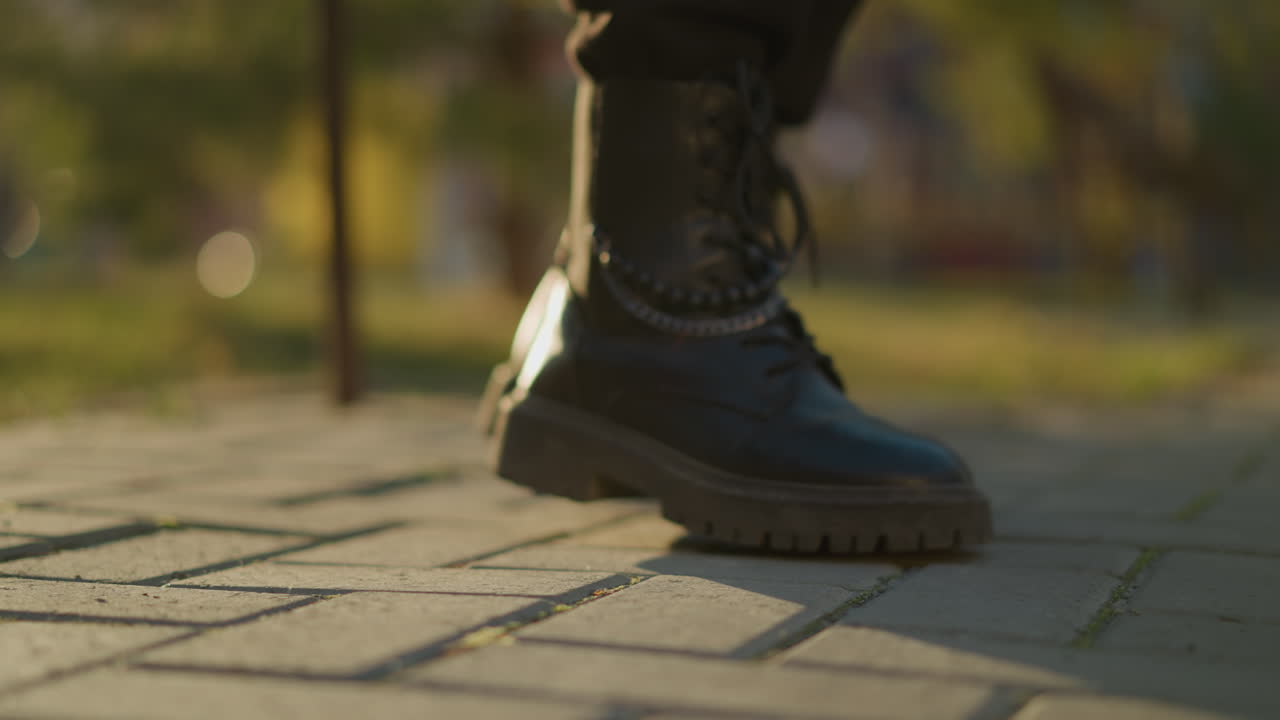 una toma de primer plano de botas negras pisando un camino pavimentado en un parque iluminado por el sol. el enfoque está en las botas, mostrando los detalles de los cordones y las suelas robustas. el fondo está suavemente borroso