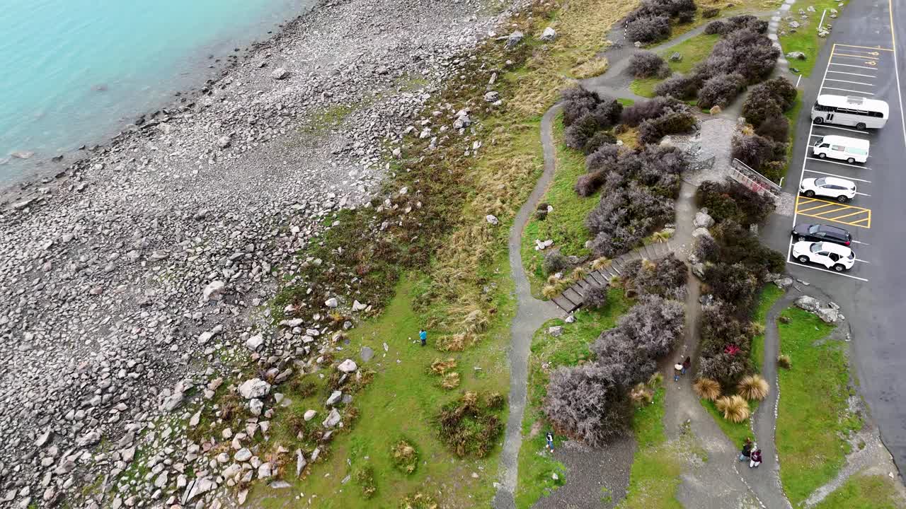Drone footage captures Lake Tekapo's rocky shoreline and adjacent parking area under natural daylight, showcasing the serene lakeside environment