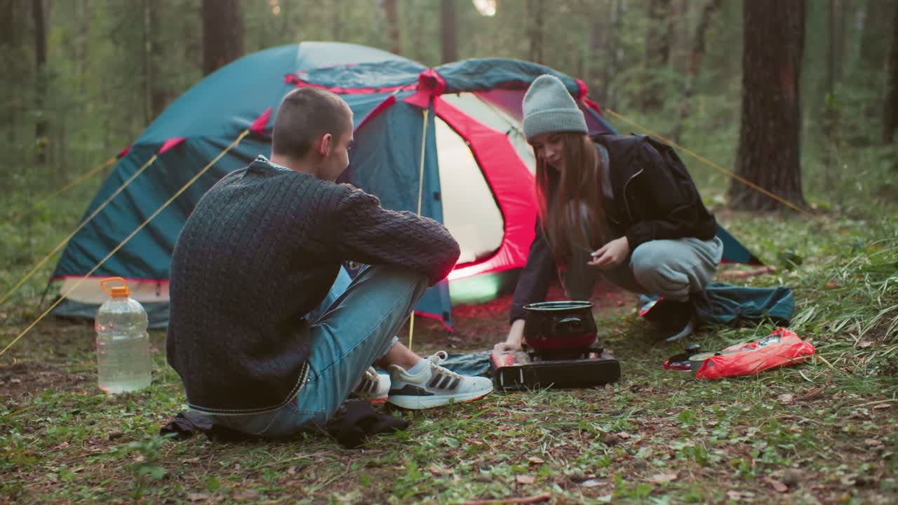 young couple spending time in forest camping as woman prepares meal using portable stove while man sits nearby smiling with tent set up in background