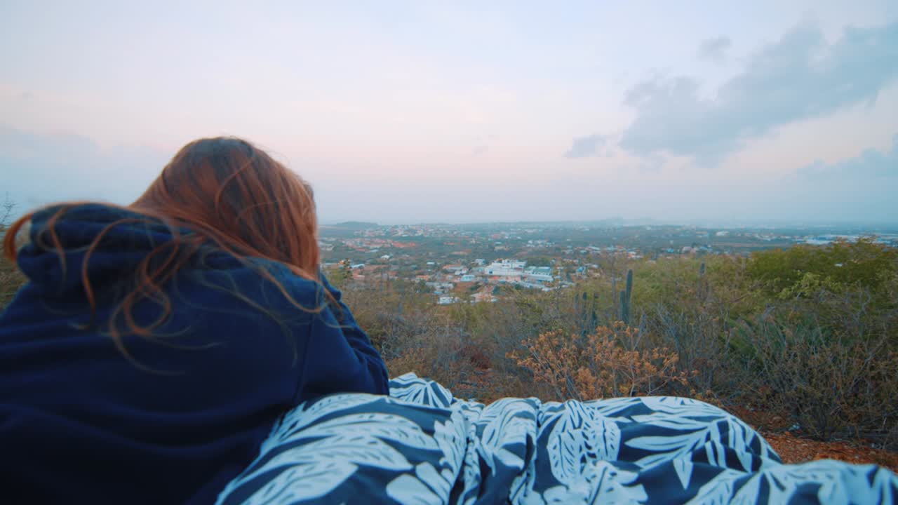 Young woman in hoodie watching sunrise and laying on blanket at viewpoint