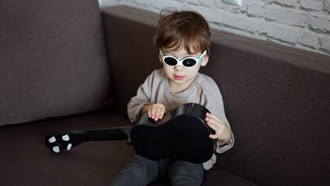 funny little kid wearing sunglasses sitting on the sofa. cute baby boy is focused on the small guitar in his hands.
