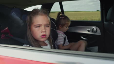 Cheerful-caucasian-girl-of-8-years-looking-out-of-the-car-window-while-car-trip-and-her-sister-in-the-background.