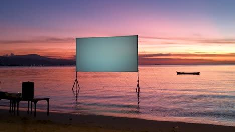 Outdoor-cinema-movie-screen-during-beautiful-sunset-reflecting-colourful-pink-and-orange-sunlight-over-ocean-on-tropical-island