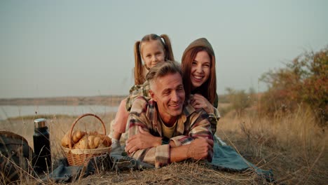 Porträt-Einer-Glücklichen-Familie-Bei-Einem-Sommerpicknick-Außerhalb-Der-Stadt.-Ein-Glücklicher-Mann-Mittleren-Alters-Mit-Grauem-Haar-In-Einem-Karierten-Braunen-Hemd-Liegt-Auf-Einer-Matte-Auf-Seinem-Bauch.-Auf-Ihm-Liegen-Seine-Frau-Und-Seine-Kleine-Tochter,-Während-Sie-Bei-Einem-Picknick-Im-Sommer-Außerhalb-Der-Stadt-Entspannen.