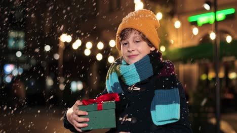 portrait of happy cute boy holding christmas present and smiling at camera on the street while it‚äôs snowing in christmas