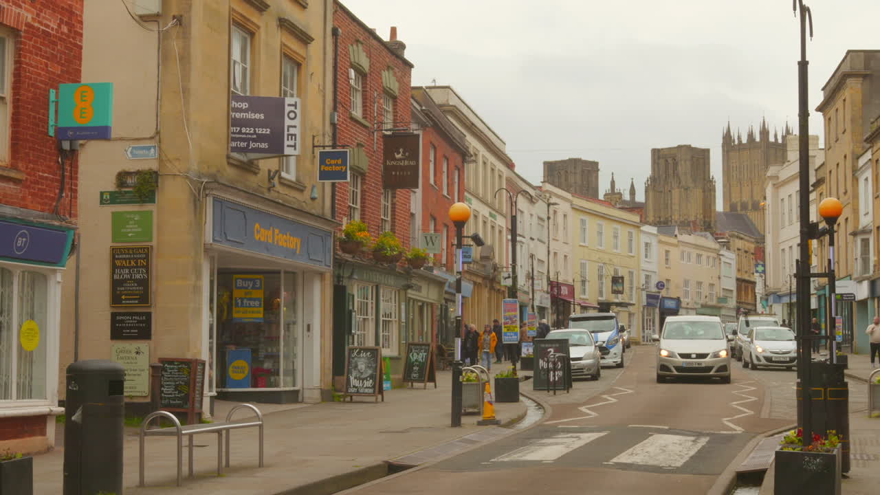Wells, London comes alive with pedestrians strolling and cars navigating the busy streets. The historic buildings tower in the background against a cloudy sky.