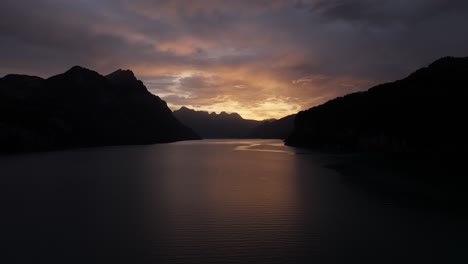 drone slowly pulls back from walensee lake, surrounded by dark mountains as morning sunrise casts orange rays on clouds, breathtaking scene in wessen, quinten, switzerland.