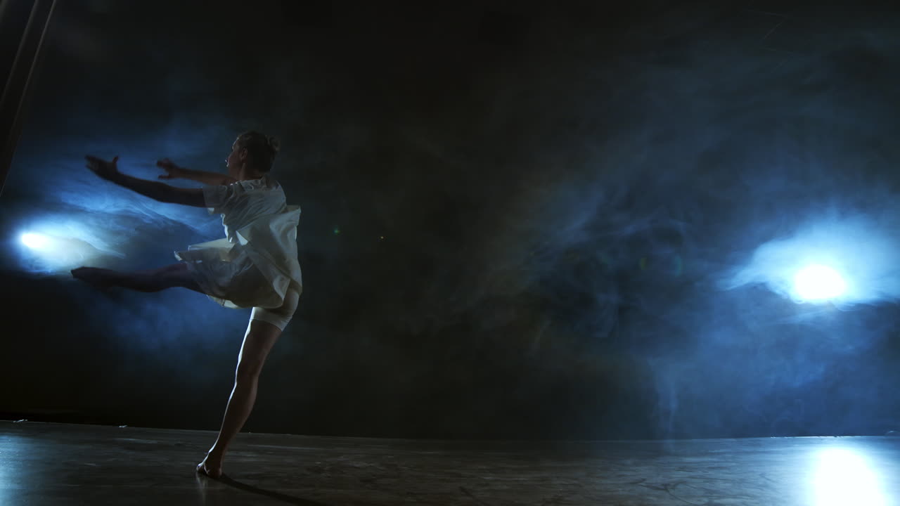 Modern dance woman in a white dress dances a modern ballet jumps on the stage with smoke in the blue spotlights.