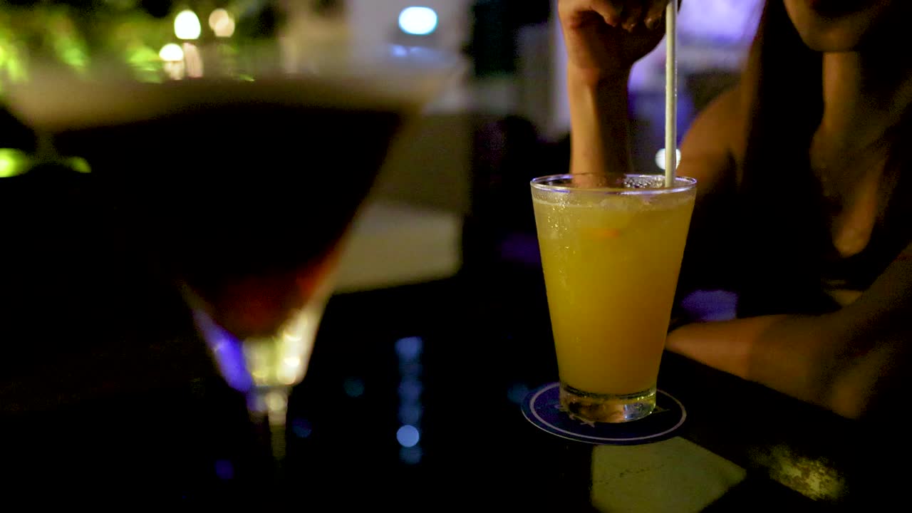 Woman stirs orange cocktail at night, bokeh lights, intimate bar, shallow depth of field