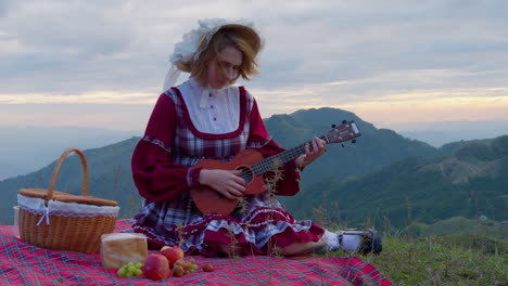 young blonde female model in old fashioned traditional clothing having lunch alone in mountains landscape while playing string guitar instrument historical reproduction