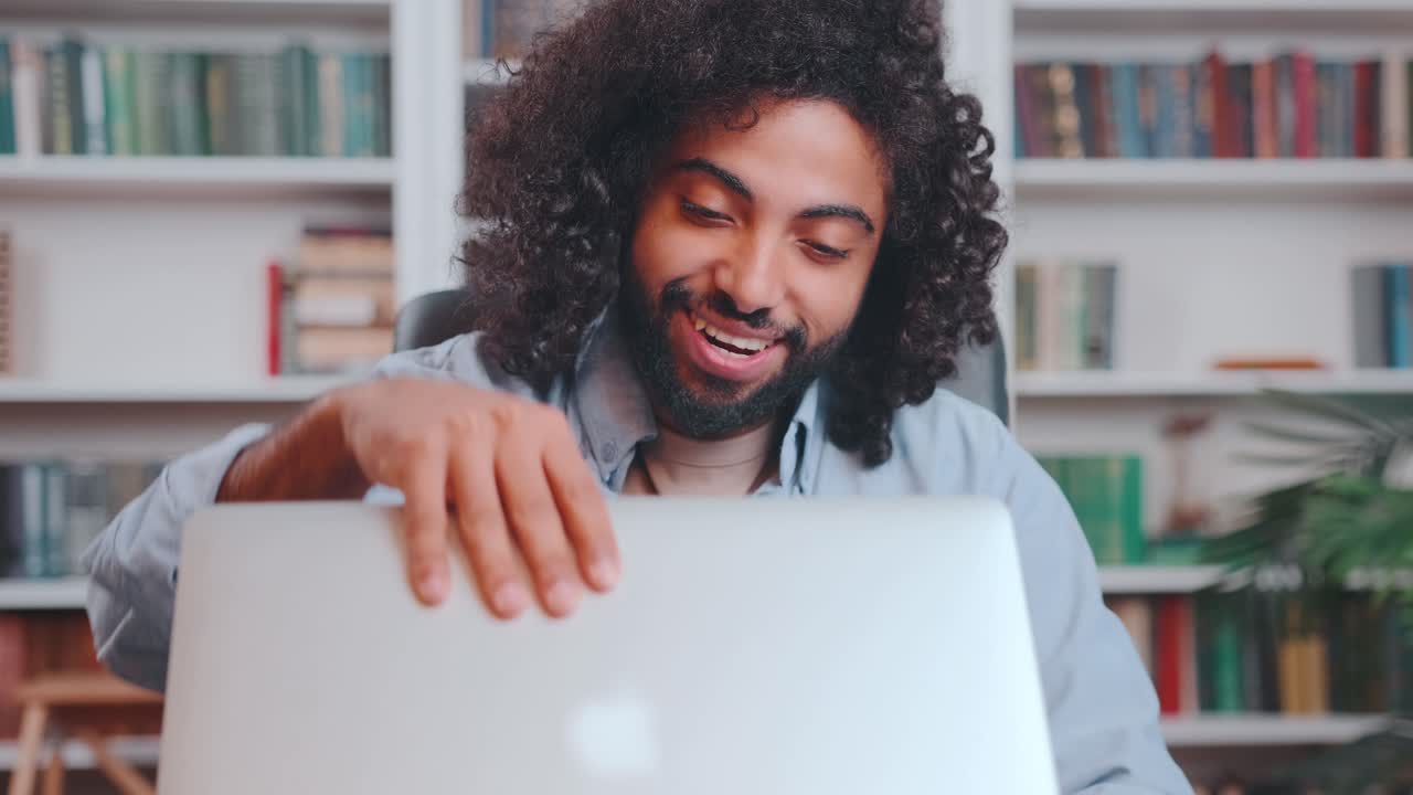 Giovane e felice freelancer arabo apre il portatile e digita, seduto nel suo ufficio domestico.