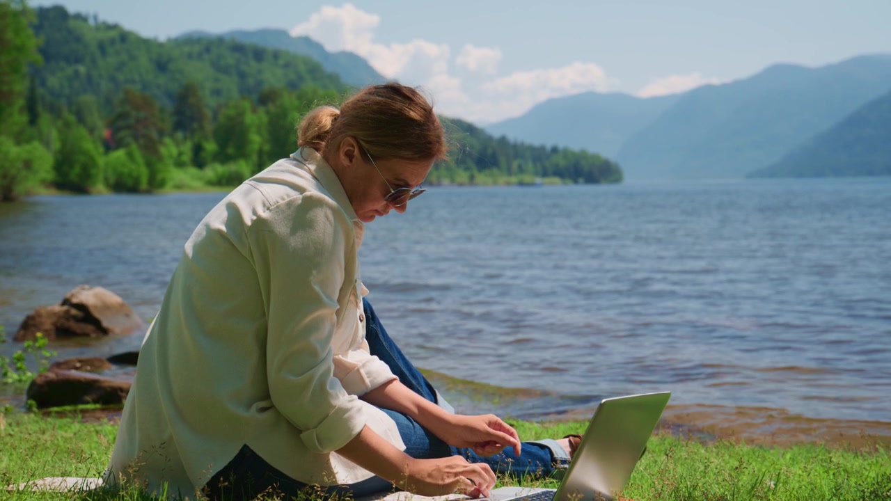 Wanita bekerja di laptop di tepi danau yang tenang. Dia fokus penuh pada layar. Gunung dan pepohonan membingkai air yang tenang. Sempurna untuk kerja jarak jauh atau tema perjalanan. Ideal untuk pemasaran gaya hidup nomaden digital.
