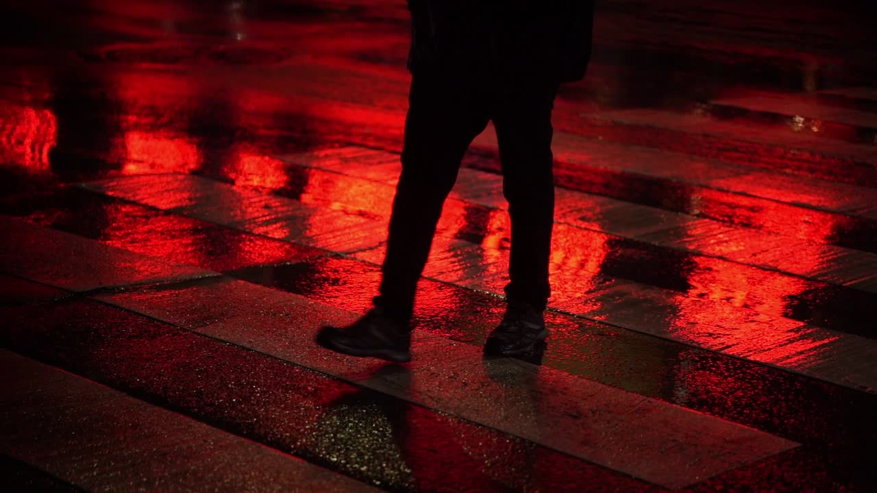 Person walks slowly on wet road crosswalk with red light reflecting on ground