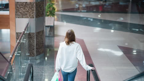 back view of woman stepping off moving escalator in spacious, brightly lit shopping mall with modern architecture, glossy floor reflecting surrounding environment, and showcasing sleek design