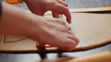 close-up of baker hands pressing orange shape cutter into smooth brown dough, wooden rolling pin and other baking tools rest nearby on textured countertop