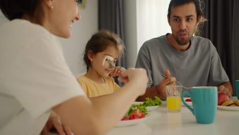 Un-Hombre-Moreno-Con-Barba-Incipiente-Desayuna-Con-Su-Esposa,-Que-Lleva-Una-Camiseta-Blanca,-Y-Su-Pequeña-Hija,-Que-Lleva-Un-Vestido-Amarillo,-En-La-Mesa-Familiar-Por-La-Mañana-En-Un-Apartamento-Moderno.