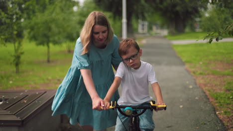 a mother assists her son as he unhappily rides his bicycle along a park walkway