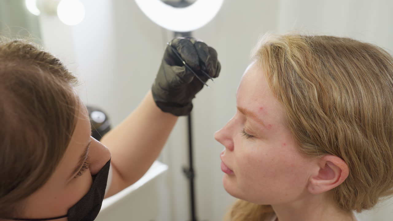 Beautician wearing gloves and face mask gently removes excessive brow hair from client using picker and brush in well-lit salon, focusing precisely on shaping client s eyebrow with professional care