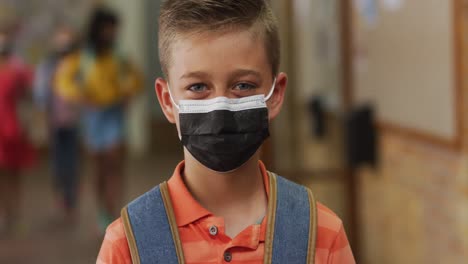 portrait of caucasian schoolboy wearing face mask, standing in corridor looking at camera