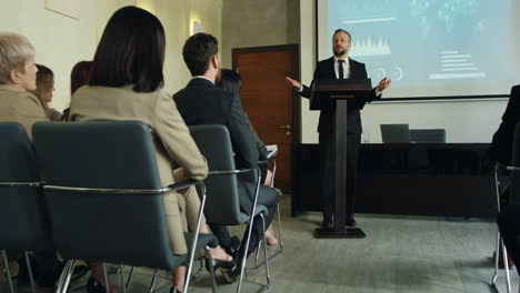 camera zoom in on caucasian businessman talking on a podium in a conference room and showing some charts and graphics on the big screen