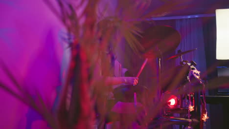 close up of decorative plant in foreground partially obscuring young girl playing electronic drums in dimly lit studio with purple and red lights, monitor screen glowing in background