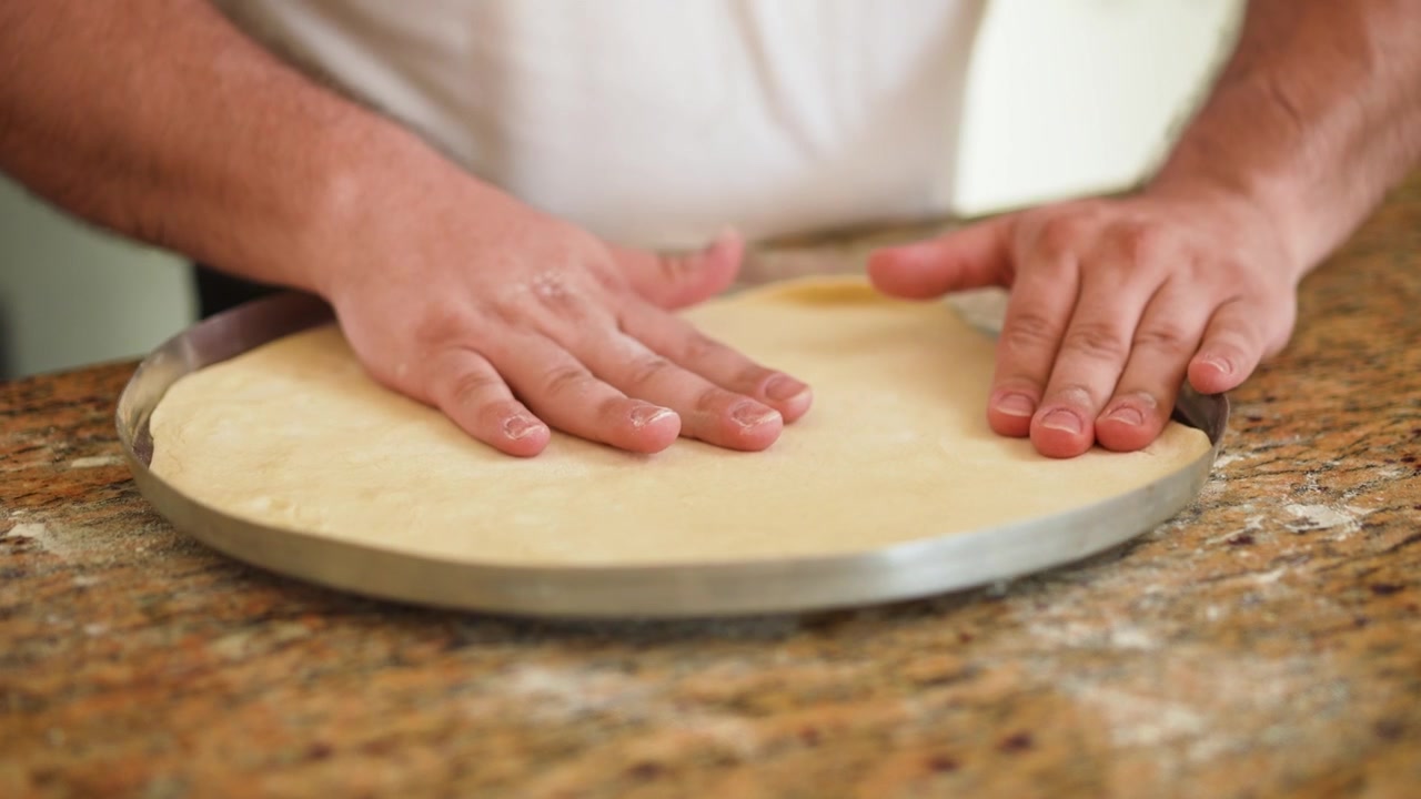 Un momento culinario catturato mentre le mani modellano abilmente l'impasto fresco della pizza in una teglia, evidenziando tecniche di cottura rustiche e la consistenza della crosta non cotta.