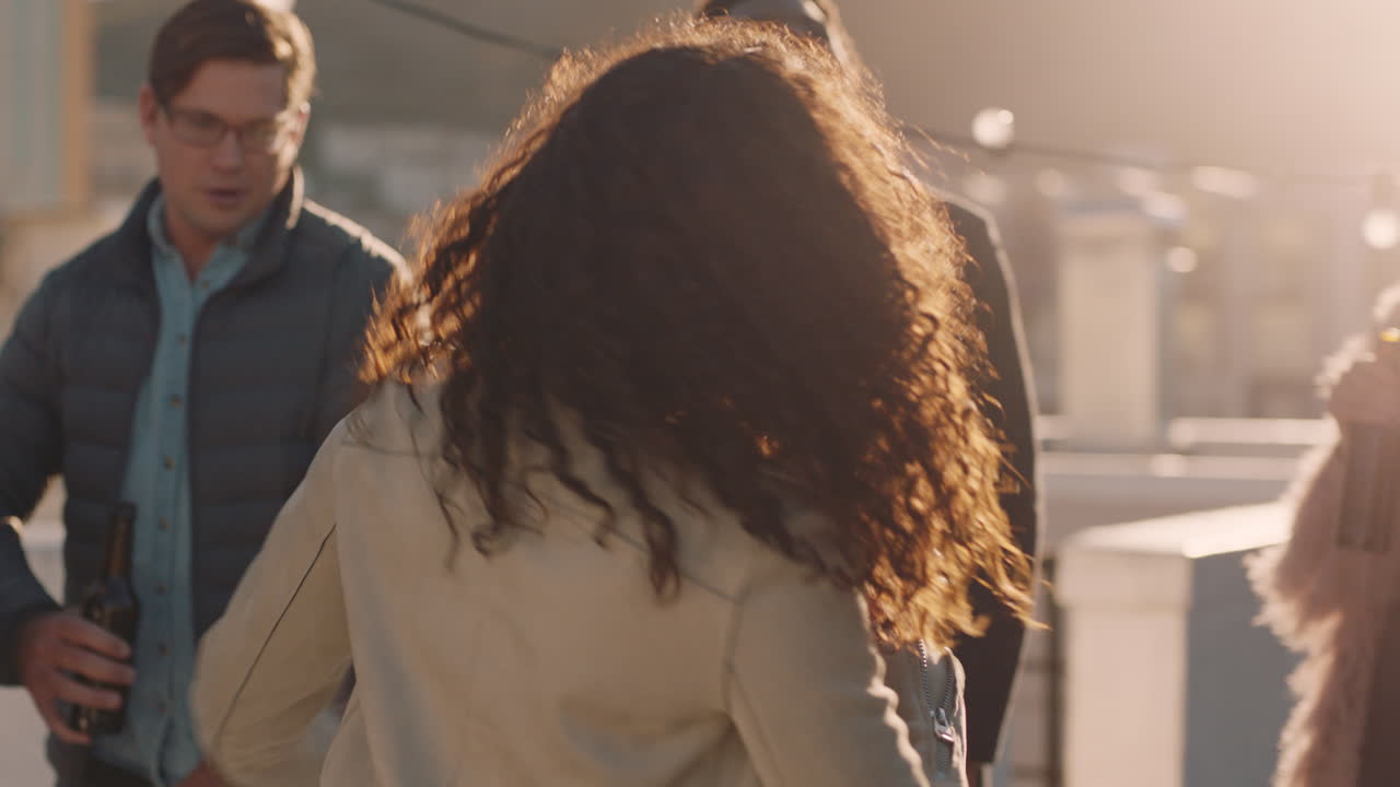 젊은 여성이 춤을 추고 다양한 친구들의 그룹이 함께 어울려서 해가 지는 날 옥상 파티를 즐기고 알코올을 마시고 주말 축하를 즐기고 있습니다.