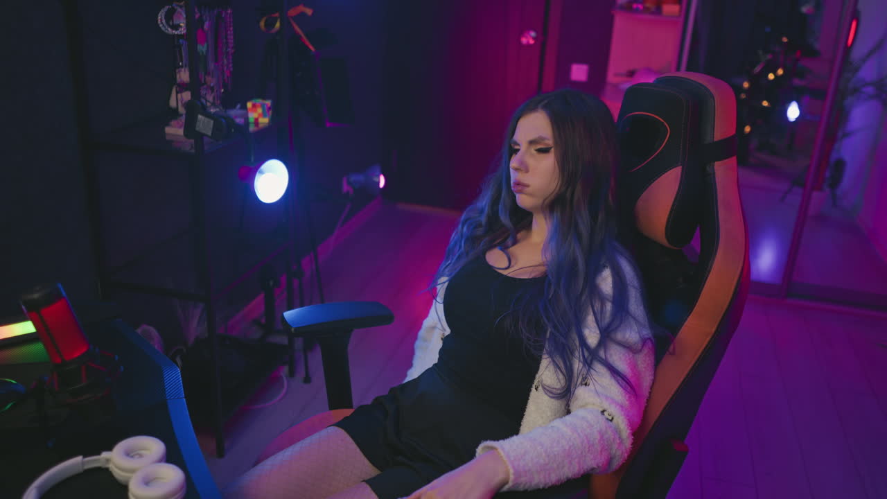 frustrated young woman leans back in gaming chair to rest after working on digital task with headset, condenser microphone, and moody lighting setup in tech workspace featuring mirror reflections