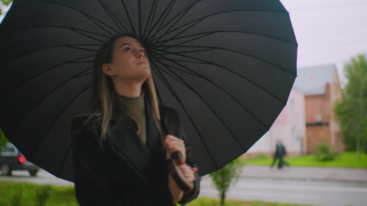 Person in black coat holds large folded umbrella obscuring face while standing outdoors on urban street with blurred cars and trees in background, creating dramatic moody rainy day atmosphere
