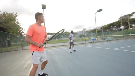 Two-diverse-male-friends-playing-doubles-returning-ball-on-outdoor-tennis-court-in-slow-motion