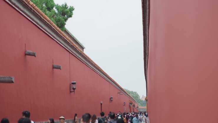 Stretto corridoio con pareti rosse e folla asiatica, pellegrini e guide turistiche che si fanno strada in una fitta processione, base in pietra antica e grondaie piastrellate che incorniciano una prospettiva stretta, dettaglio architettonico tranquillo sotto un cielo grigio