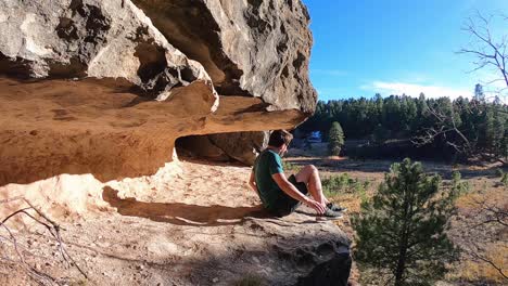 a man climbs up on a rock shelf overlooking a forest, sits down with his legs hanging off the side of the cliff's edge